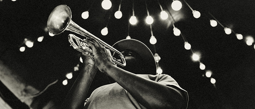 Black and white photograph of musician playing trumpet under string lights, capturing the intimate atmosphere and musical heritage central to blues origins and development