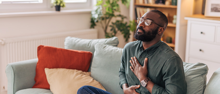 Man sitting on sofa practicing mindfulness with closed eyes and hands on chest