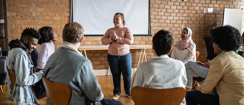 In a community centre room, a woman in jeans and a pink sweater is smiling and speaking to a small seated circle of men and women from all cultures