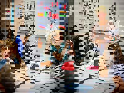 A smiling teacher in a light pink sweater holds papers while standing in a classroom, engaging with diverse students seated around her.  Students have laptops open, and one student in a teal shirt appears to be actively participating in the discussion, creating a welcoming multicultural learning environment.