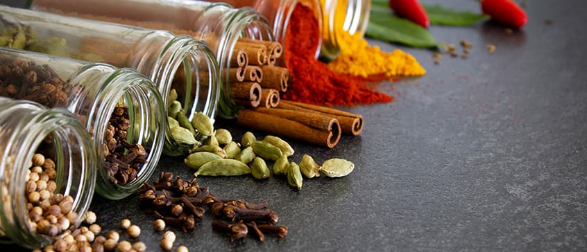 An overhead shot displays an array of colorful spices on a dark gray surface. Clear glass jars are tipped over, spilling whole white peppercorns, cloves, green cardamom pods, and cinnamon sticks. Piles of red and yellow powdered spices are also visible, along with loose cloves and cardamom pods. In the background, out-of-focus red chili peppers and green leaves add to the vibrant composition.