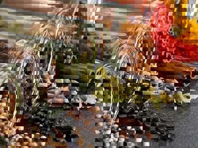 An overhead shot displays an array of colorful spices on a dark gray surface. Clear glass jars are tipped over, spilling whole white peppercorns, cloves, green cardamom pods, and cinnamon sticks. Piles of red and yellow powdered spices are also visible, along with loose cloves and cardamom pods. In the background, out-of-focus red chili peppers and green leaves add to the vibrant composition.